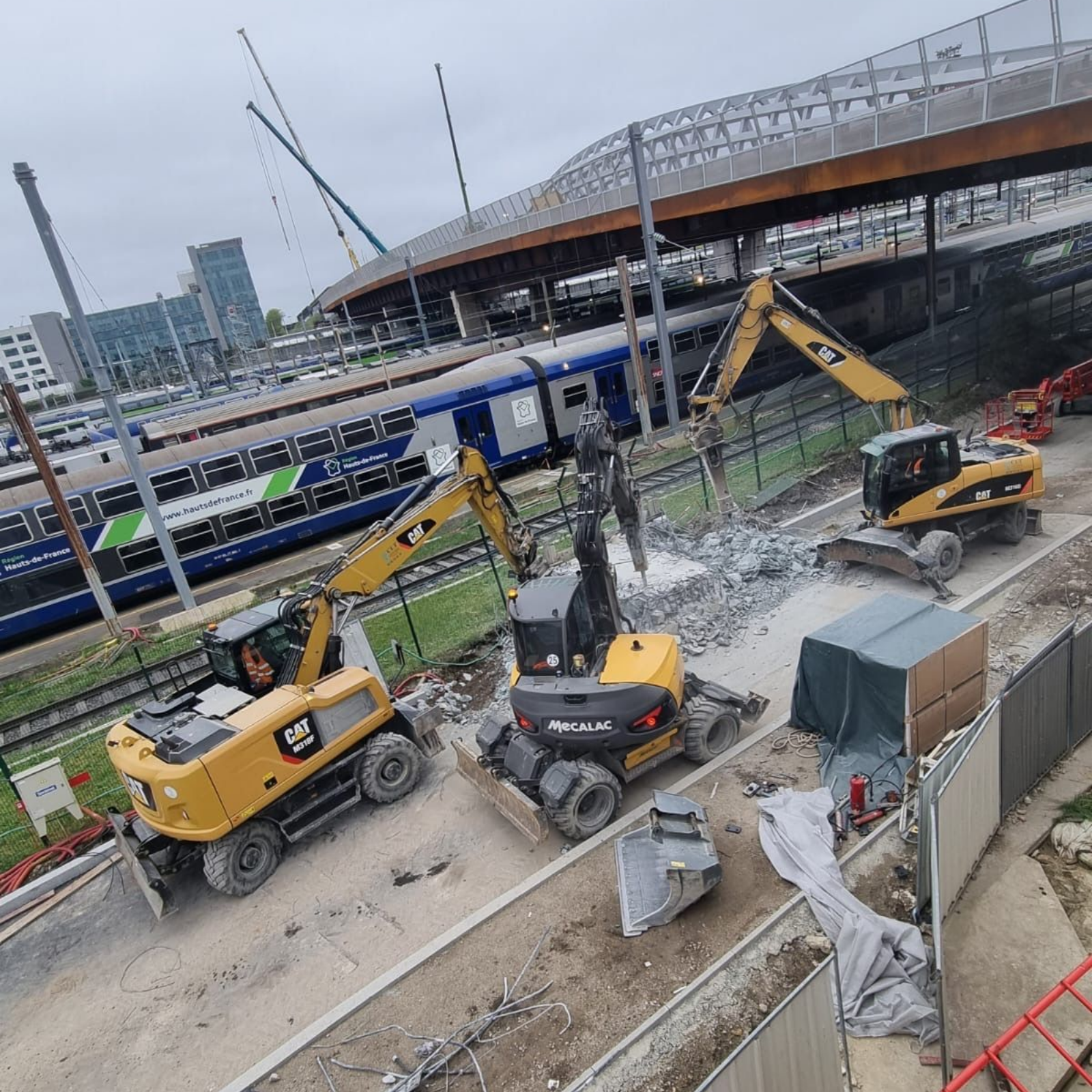 Trois excavatrices travaillent sur un chantier de construction à proximité d'une gare ; les couleurs dominantes sont le gris, le jaune et le bleu.
