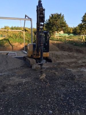 Une mini pelleteuse jaune avec tarière fore un trou dans le sol sur un chantier de construction.