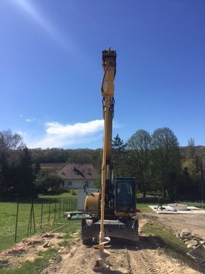 Une excavatrice jaune (pelle à pneu 20T), équipée d'une tarière, fore le sol.