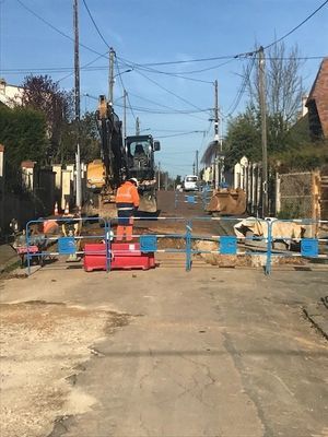 Chantier bloquant une rue, ouvriers à proximité d'engins lourds et d'une barricade, lignes électriques aériennes.