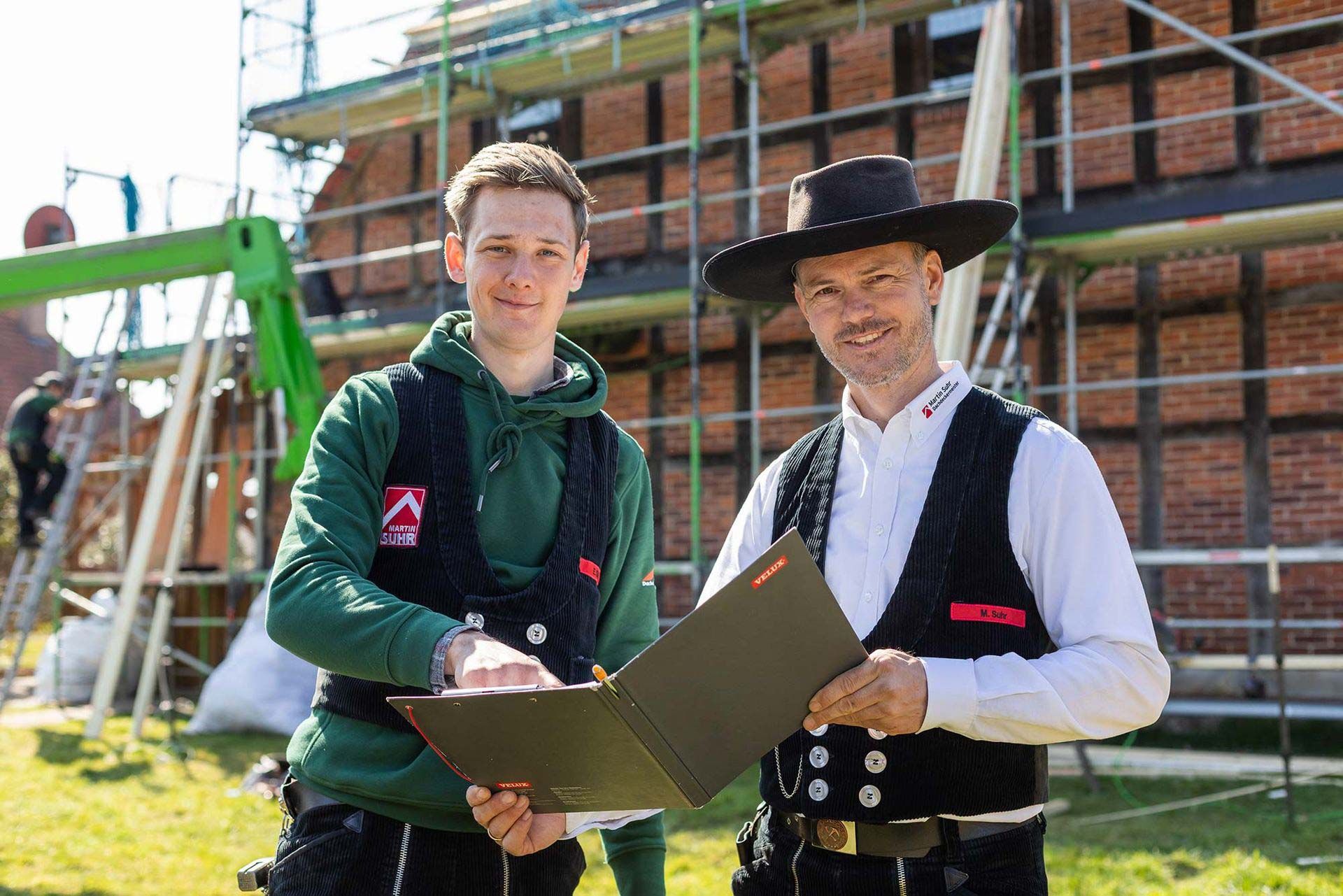 Zwei Männer stehen nebeneinander vor einem Backsteingebäude und halten ein Klemmbrett in der Hand.