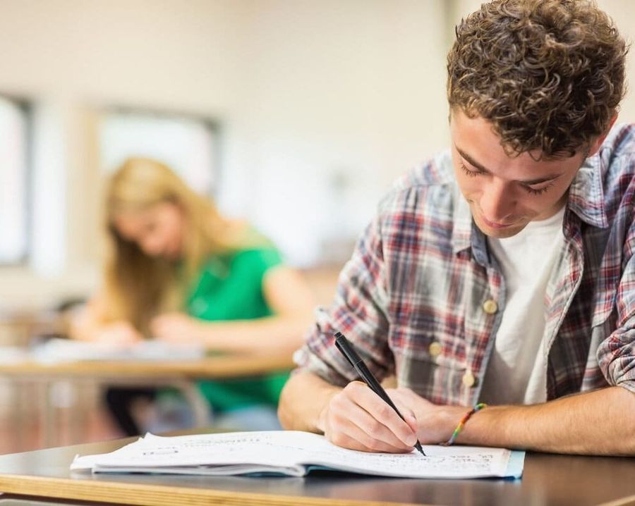 Un estudiante escribiendo en un aula, con otro estudiante borroso al fondo.