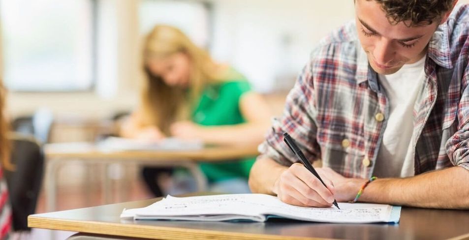 Estudiante realizando un examen en un aula, con un compañero de clase borroso al fondo.