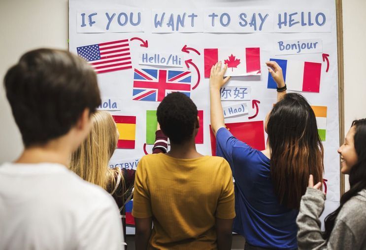 Los alumnos señalan un tablero de aprendizaje de idiomas con banderas y saludos en diferentes idiomas.