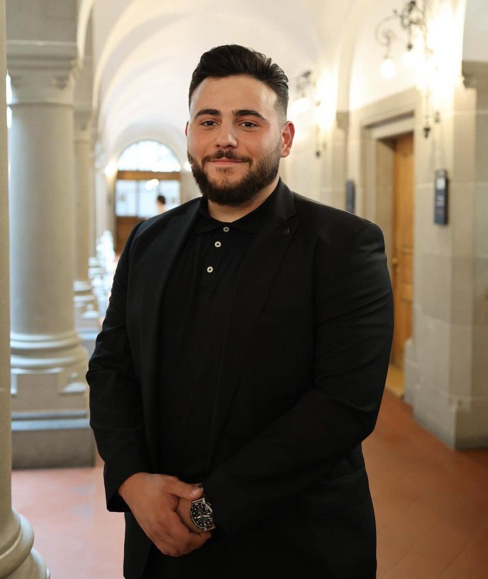 Ein Mann mit Bart steht in einem Flur mit Säulen.