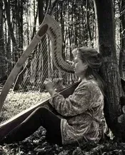 Frau spielt Harfe in einem Wald, an einen Baum gelehnt. Schwarzweißfoto.