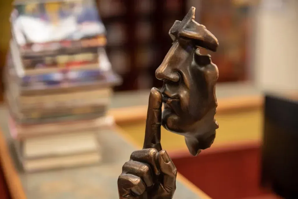 Bronzestatue eines Gesichts mit einem Finger auf den Lippen, in einer „Psst“-Geste.