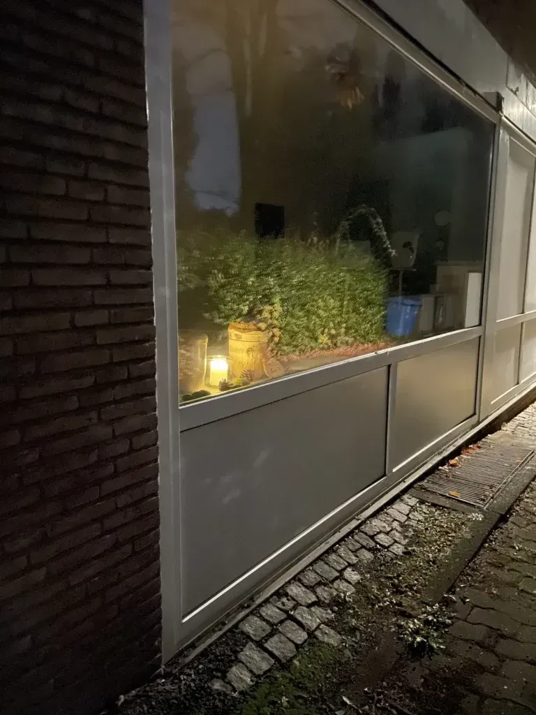 Ein durchsichtiger Plastikvorhang schützt ein Fenster. Hinter dem Vorhang sind Pflanzen und brennende Kerzen zu sehen.