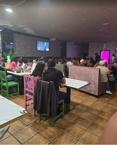 Gente sentada en el comedor de un restaurante con poca luz, televisores en las paredes y suelos de baldosas.