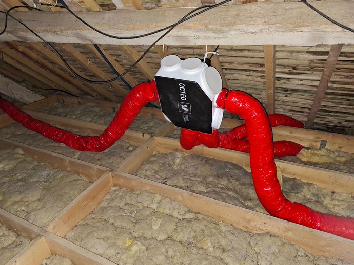 Unité de ventilation installée dans un grenier. Les conduits, l'isolation et les poutres en bois sont visibles.