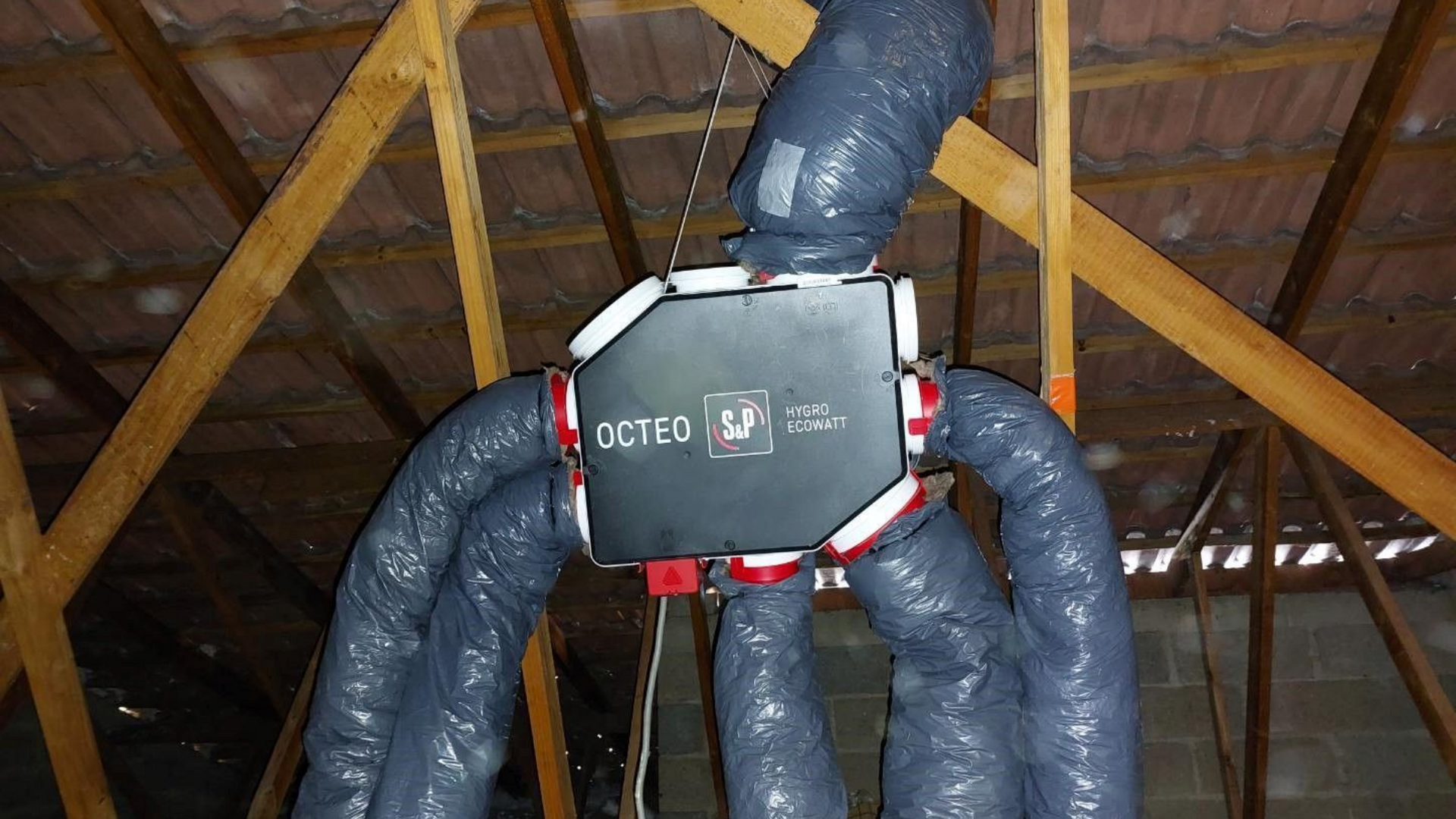 Unité de climatisation dans un grenier avec conduits isolés gris reliés à un boîtier noir. Les poutres en bois sont visibles.