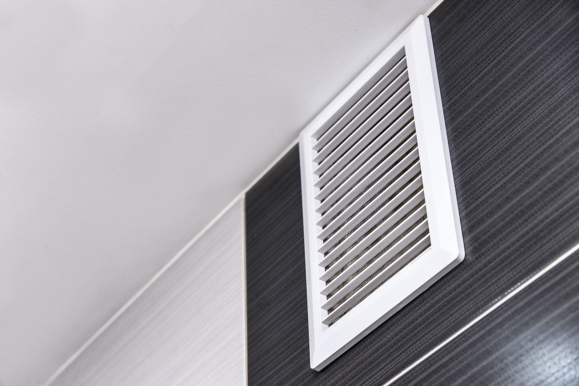 Grille d'aération blanche sur un mur carrelé noir et gris, près d'un plafond blanc.