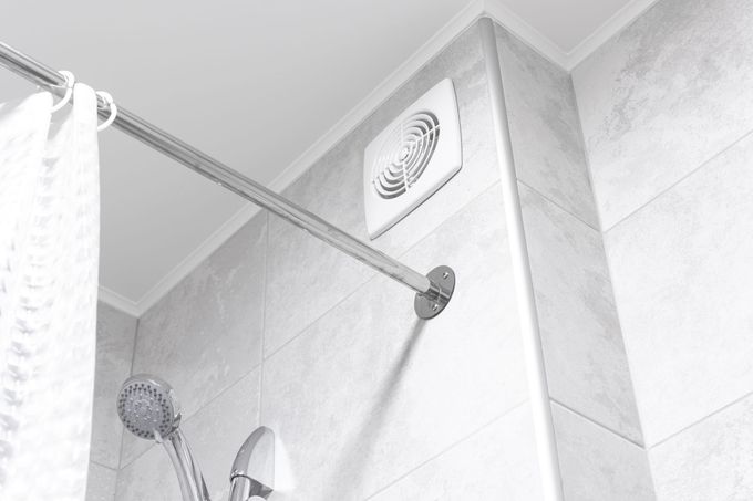Intérieur de la douche avec ventilateur, pommeau de douche, tringle à rideau et carrelage clair.