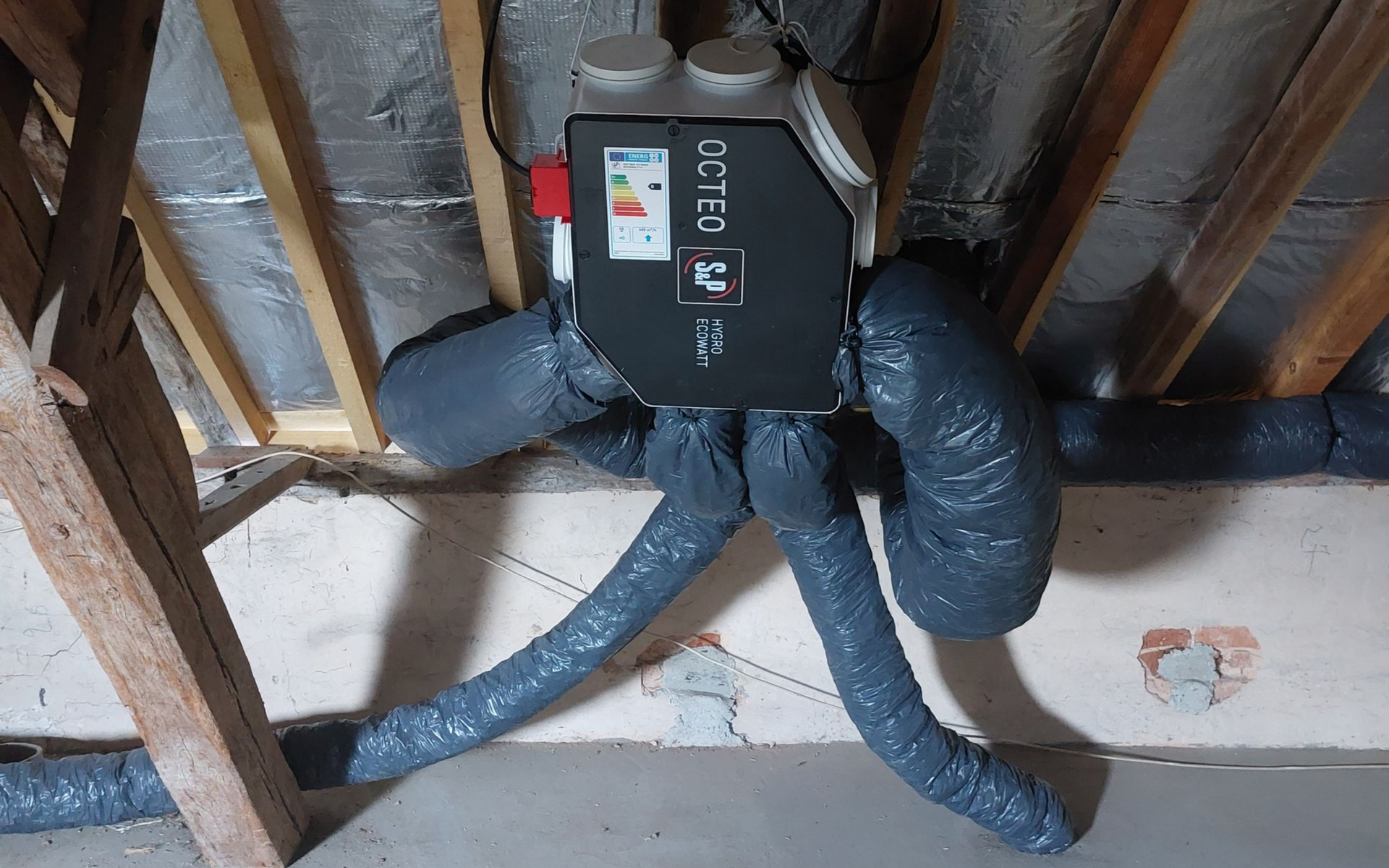 Unité de ventilation dans les combles avec conduits gris flexibles. Unité noire avec étiquette d'efficacité énergétique.