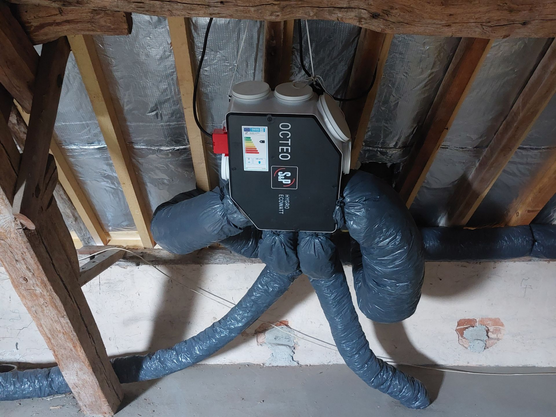 Unité de ventilation noire dans les combles, reliée à des conduits flexibles. Poutres en bois et plafond isolé visibles.