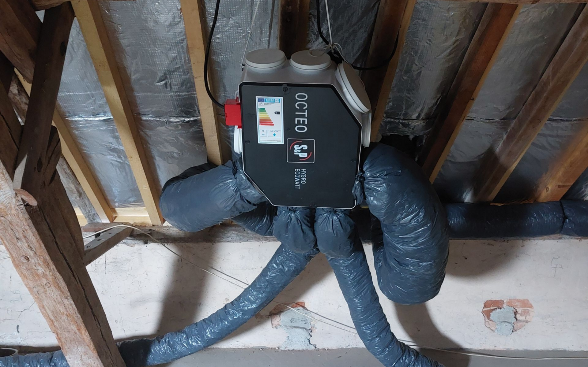 Unité de ventilation noire installée dans un grenier avec conduits isolés, poutres en bois et mur en briques.