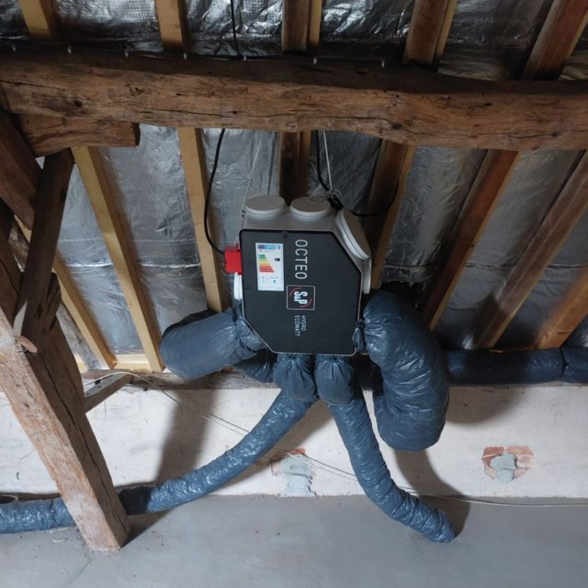 Un système de ventilation installé dans un grenier avec des conduits isolés, fixés aux poutres et chevrons en bois.