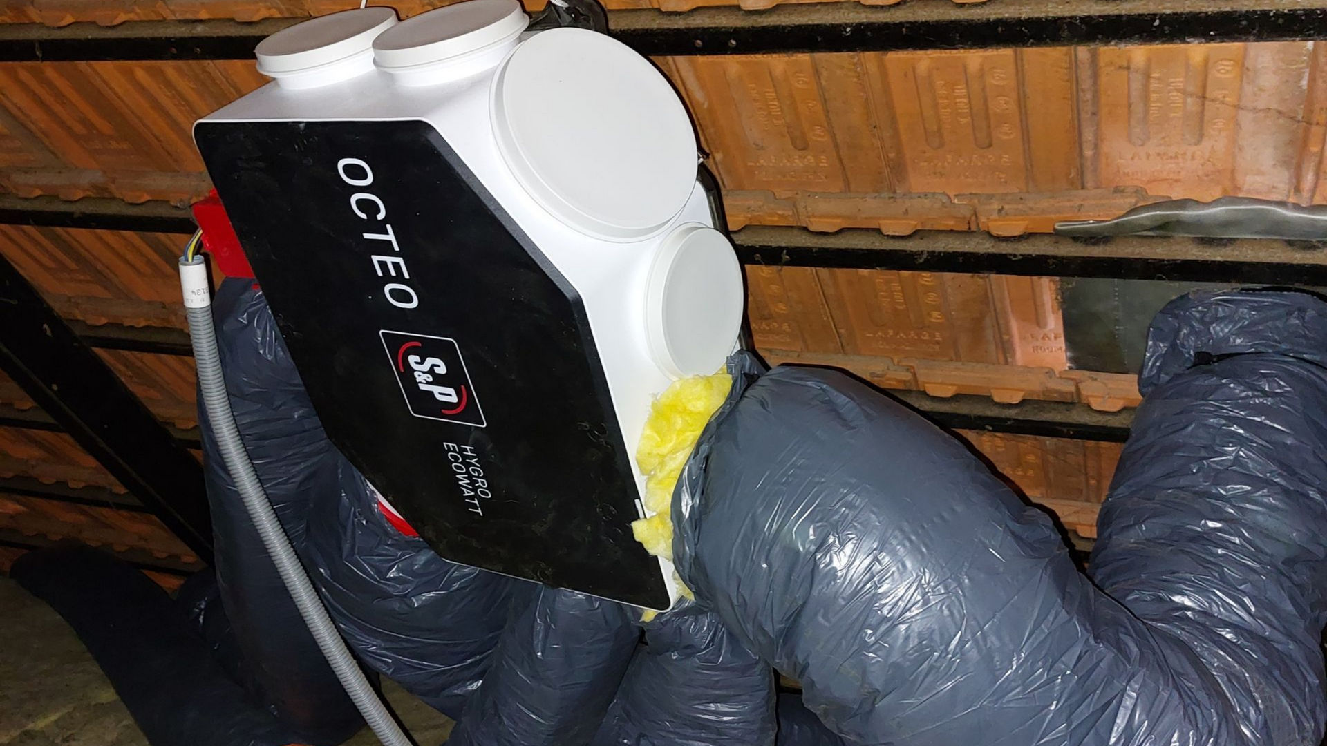 Unité de ventilation noire et blanche avec conduits dans un grenier.