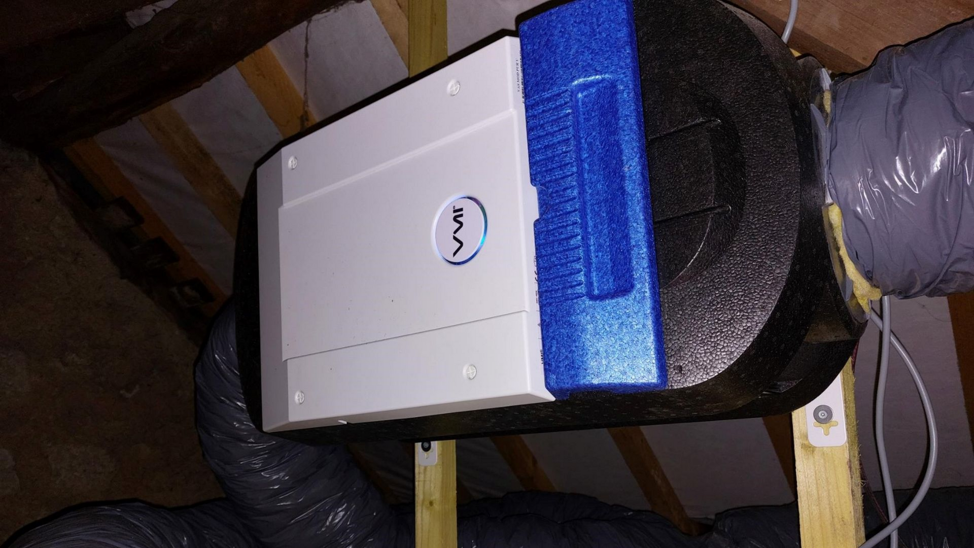 Unité de climatisation blanche et bleue installée dans un grenier, avec des conduits isolés.