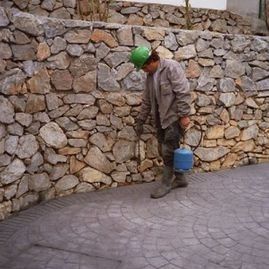 Un hombre que lleva un casco verde está parado frente a un muro de piedra sosteniendo un cubo.