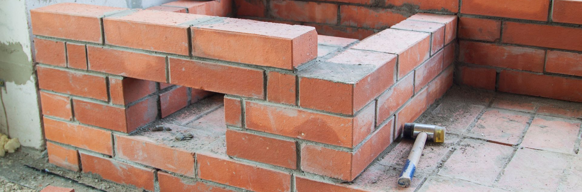 Construction en briques d'une petite cheminée extérieure, avec un marteau posé sur le sol carrelé.