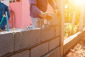 Un ouvrier du bâtiment pose des parpaings, il construit un mur. Le soleil brille de mille feux.