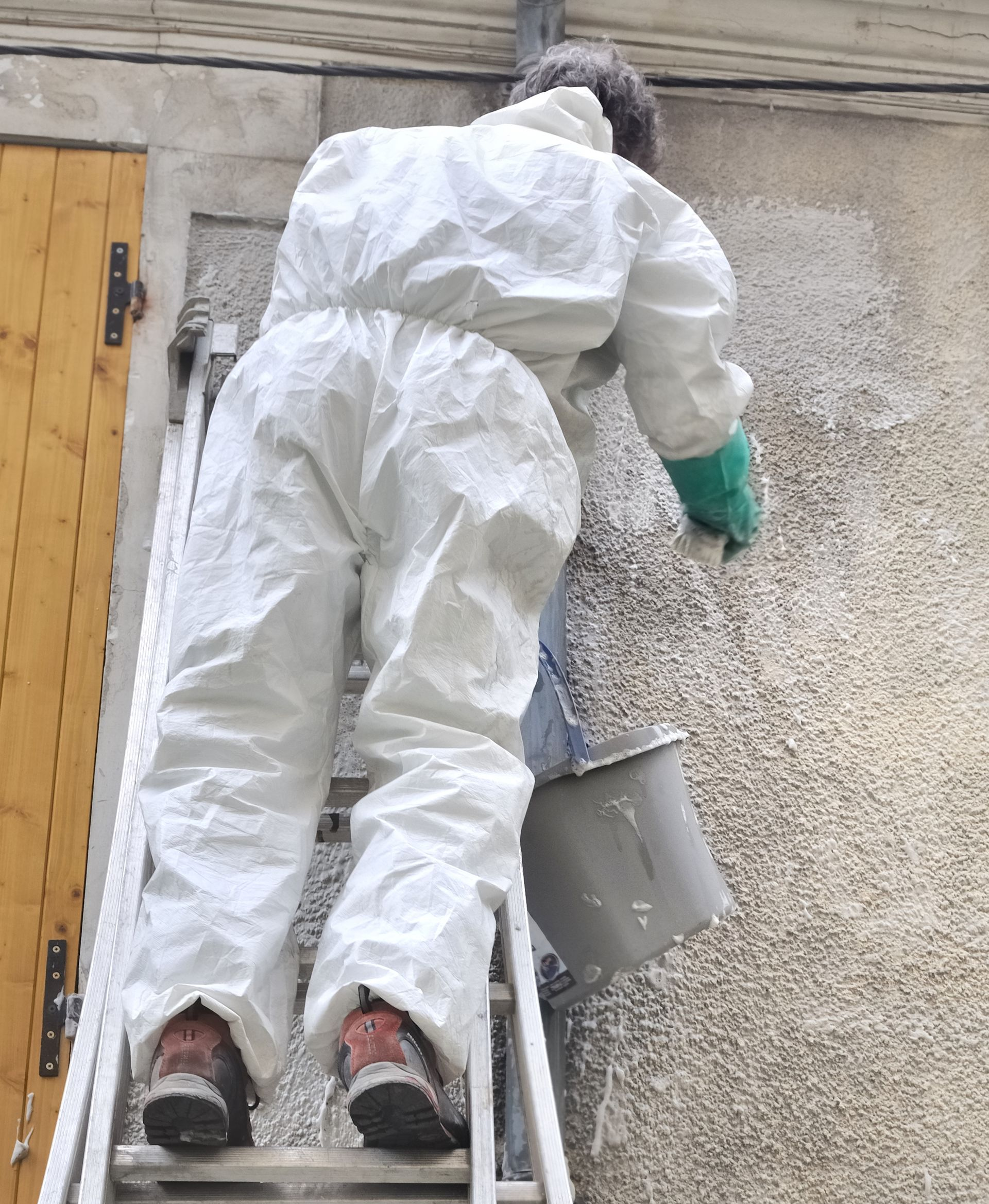 Personne en combinaison de protection en train d'enduire l'extérieur d'un bâtiment depuis une échelle, tenant un seau.