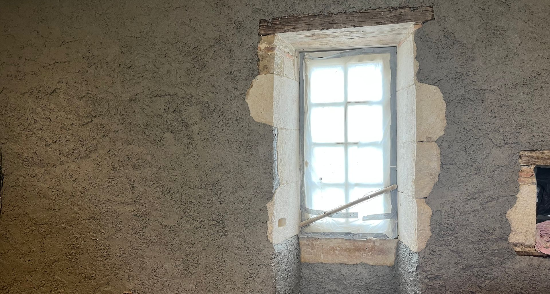 Fenêtre dans un mur de pierre patiné, avec un cadre en pierre apparente et une fenêtre usée aux rideaux blancs.