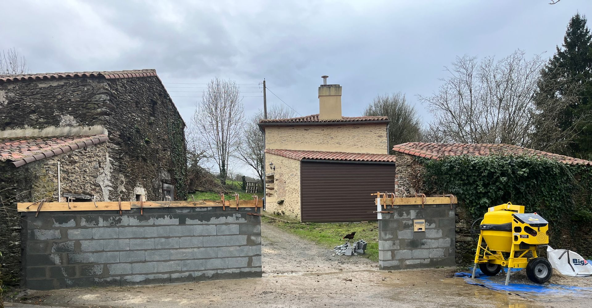 Construction d'un mur en parpaings à l'aide d'une bétonnière devant un bâtiment en pierre par temps nuageux.