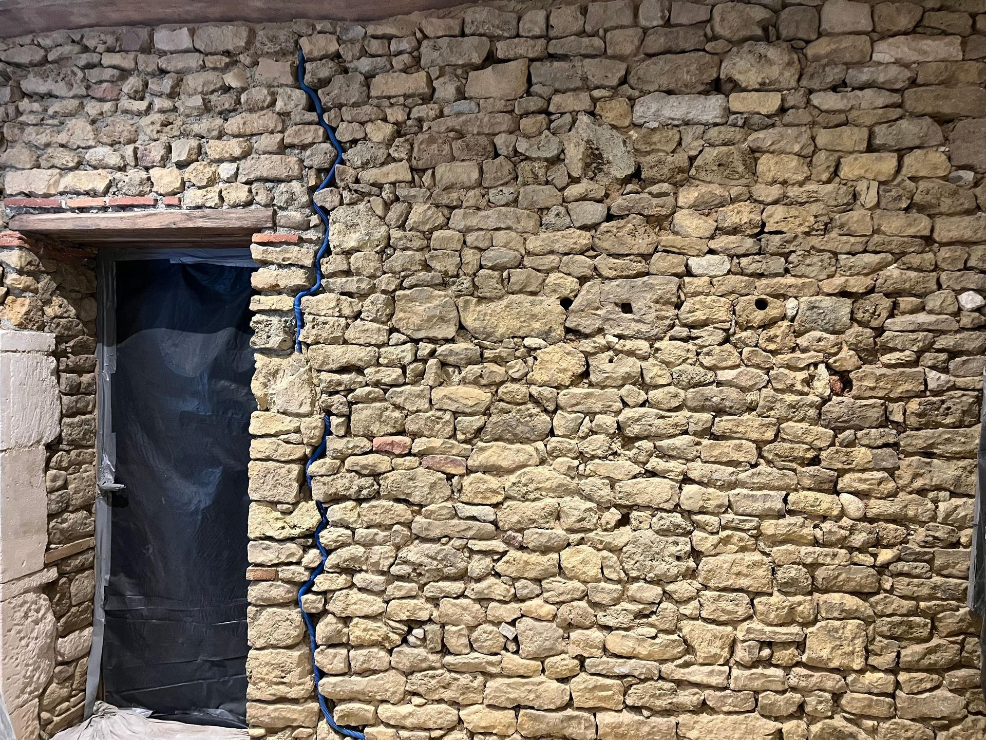 Mur en pierre avec une fissure verticale, à côté d'une ouverture de porte recouverte de plastique noir.