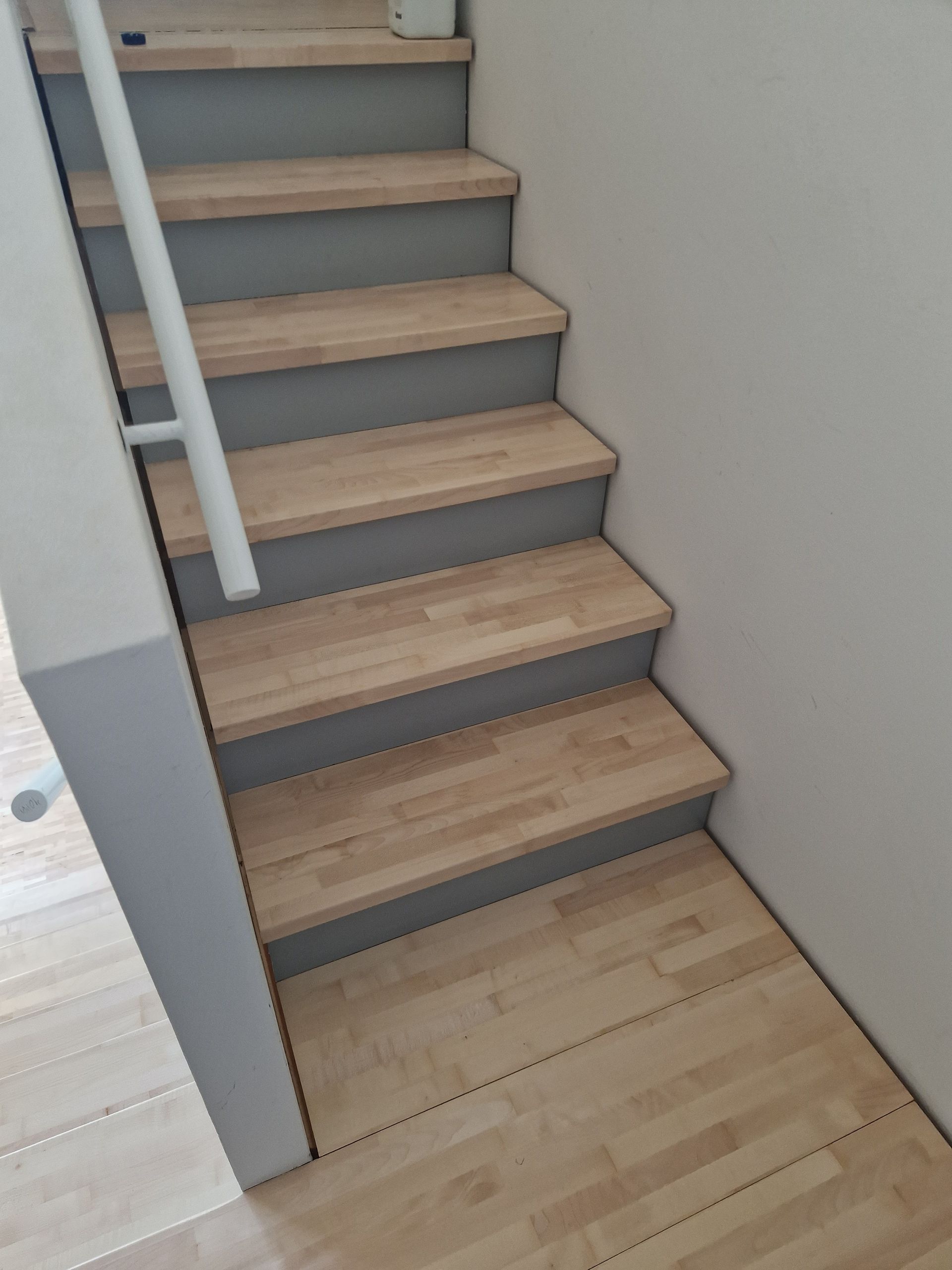 Treppe mit hellen Holzstufen und grauen Setzstufen, daneben ein weißes Geländer.