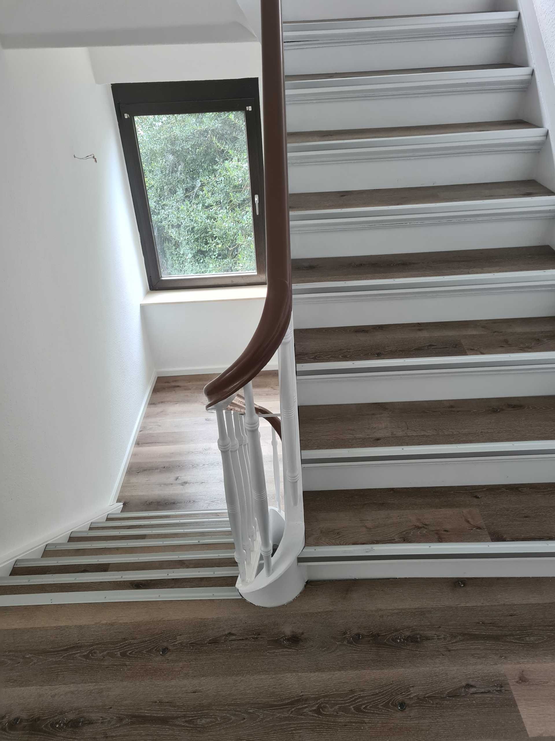 Treppe mit weißem Geländer und Stufenbelag in Holzoptik, seitlich ein großes Fenster.