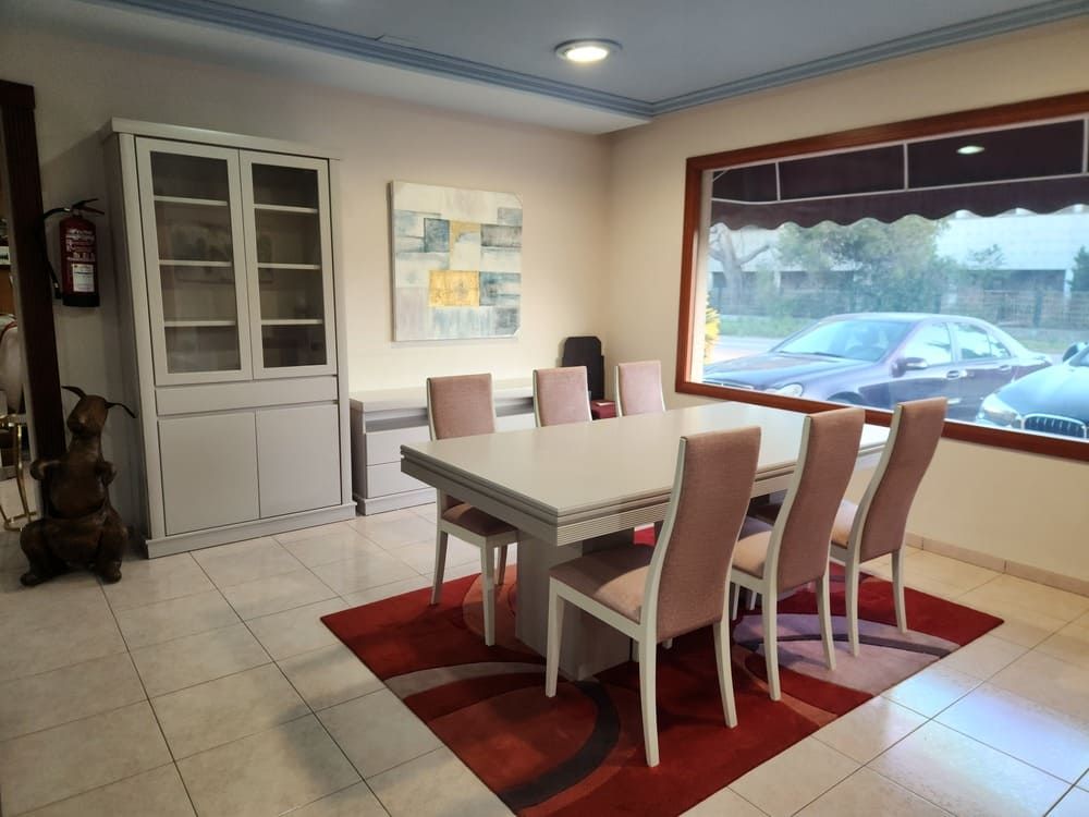 Un comedor con una mesa blanca, seis sillas rosas, una vitrina y una alfombra roja estampada sobre un suelo de baldosas.