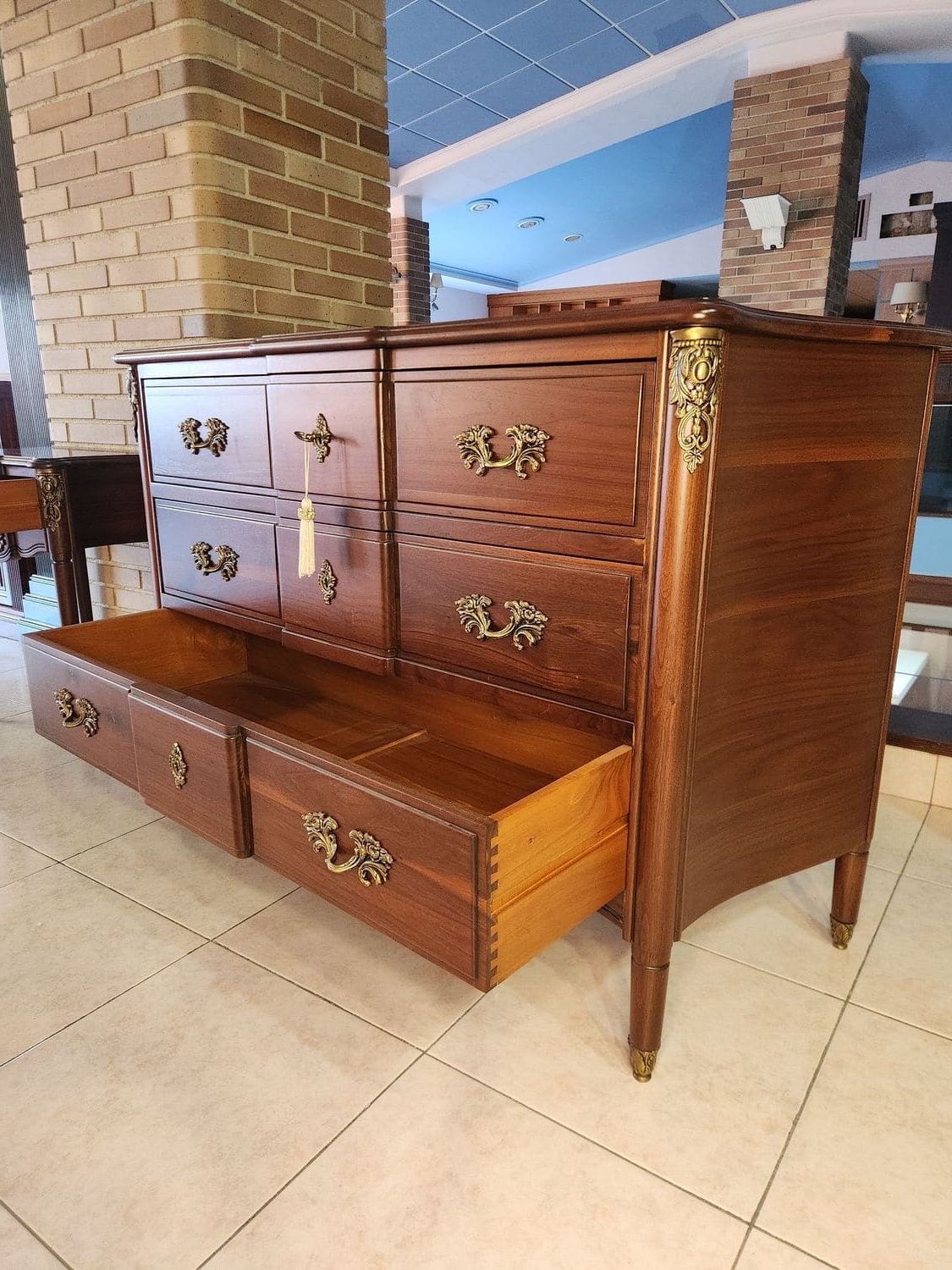 Cómoda de madera con tiradores ornamentados y un cajón abierto, sobre suelo de baldosas.