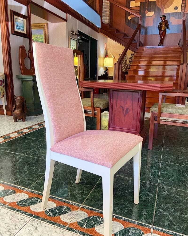 Una silla de comedor tapizada en rosa con estructura blanca descansa sobre un suelo de baldosas, frente a un conjunto de comedor de madera y una escalera.
