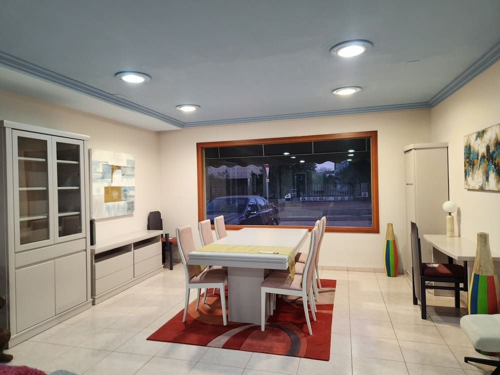 Un luminoso comedor con una mesa blanca y seis sillas sobre una alfombra roja estampada, junto a una gran ventana y armarios blancos.