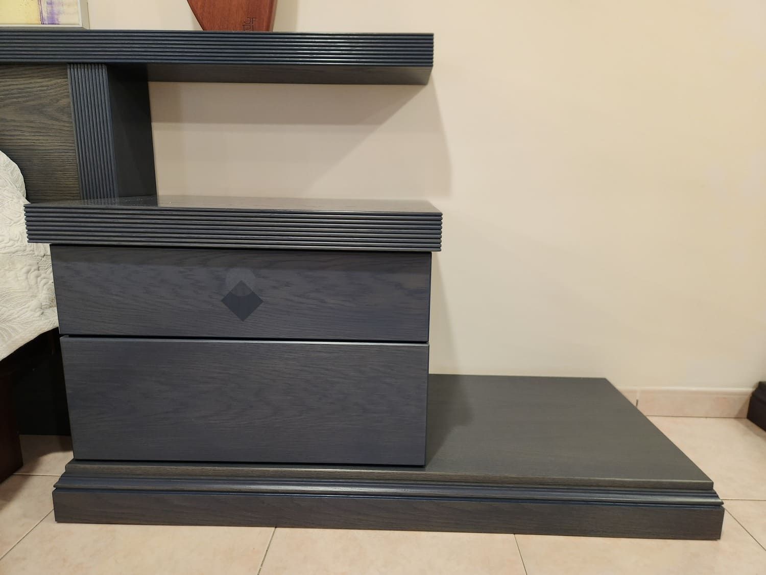 Mesita de noche gris oscuro con estante y dos cajones contra una pared beige.