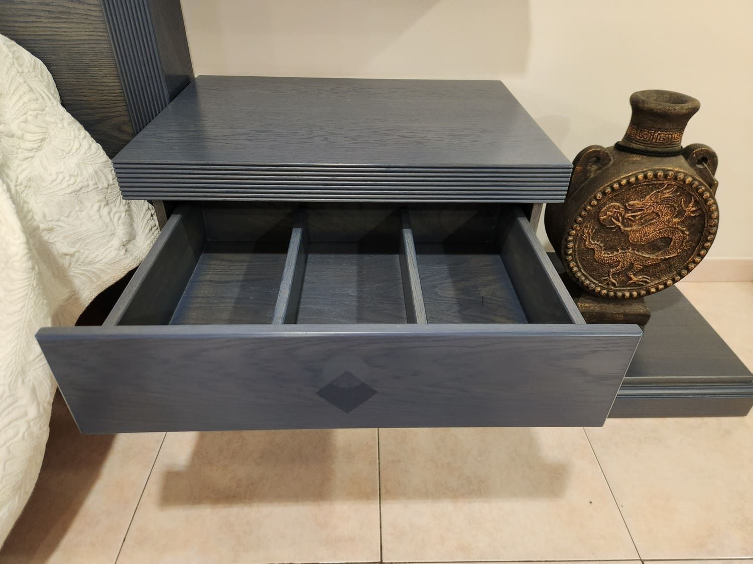 Mesita de noche azul con cajón abierto, jarrón tallado y cama parcialmente visible.