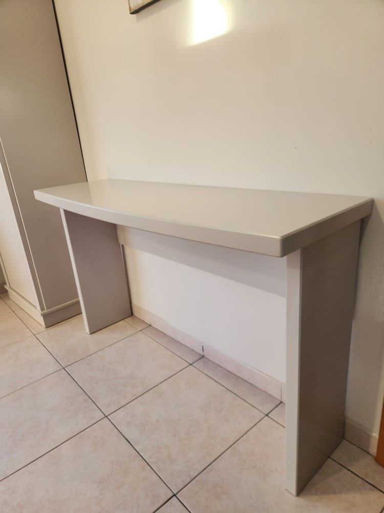 Una sencilla mesa consola de color gris claro, apoyada contra una pared blanca lisa sobre un suelo de baldosas.