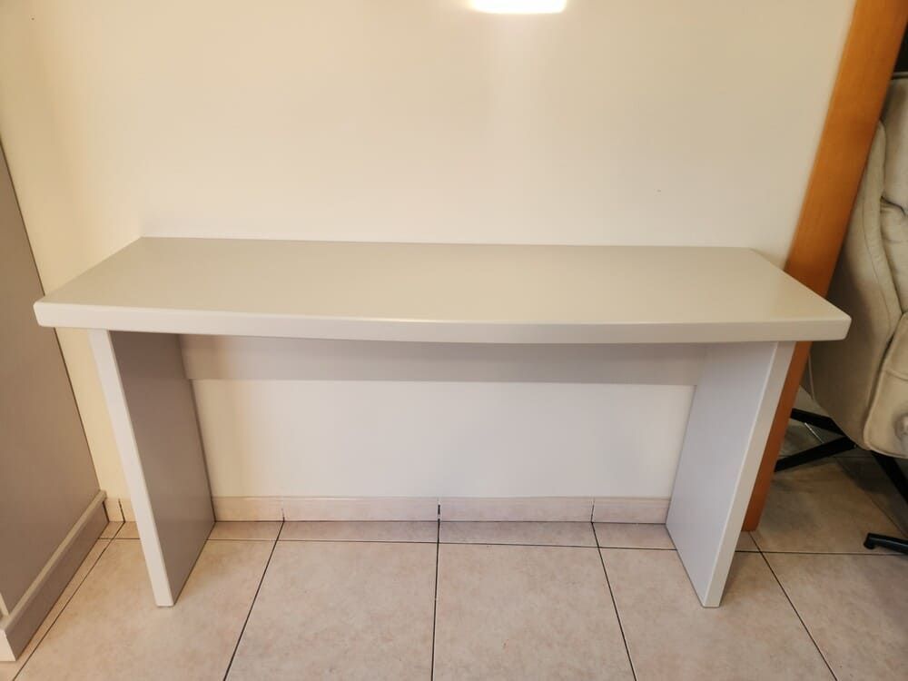 Una mesa consola minimalista de color gris claro con dos patas de estilo plano, apoyada contra una pared blanca sobre un suelo de baldosas.
