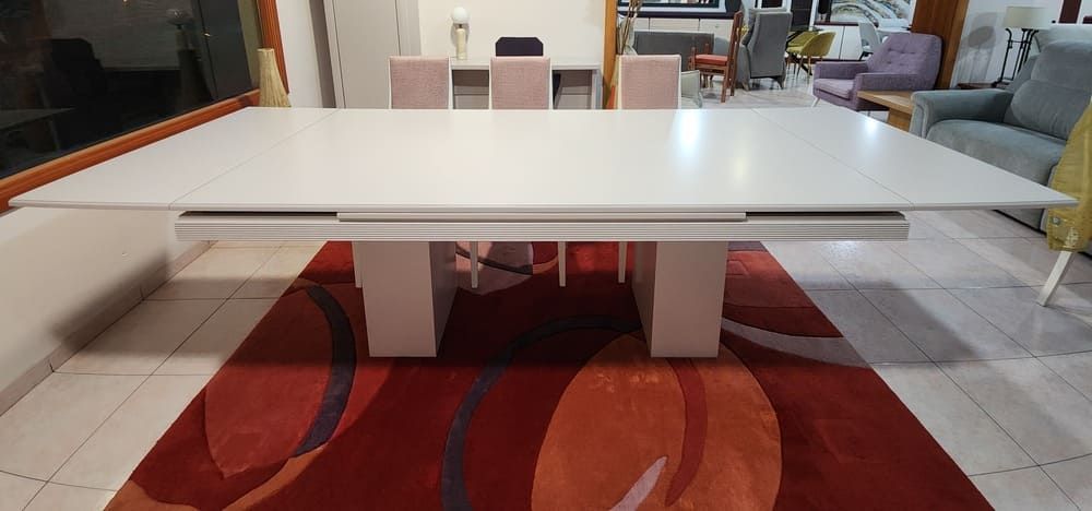 Una elegante mesa de comedor rectangular blanca con una robusta base central, colocada sobre una alfombra con estampado rojo y naranja en una sala de exposición.