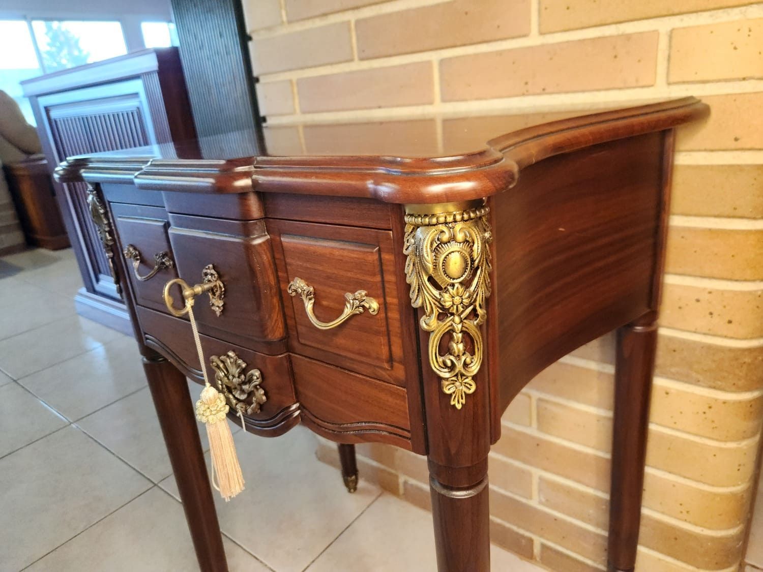 Mesita de noche de madera oscura con herrajes dorados ornamentados y tres cajones.