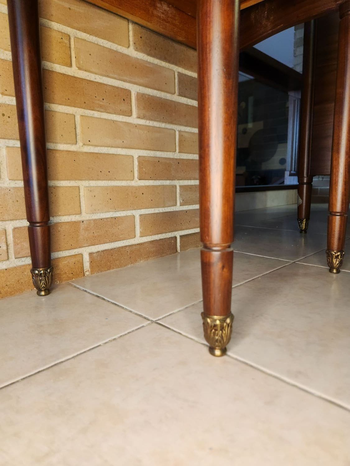 Patas de mesa de madera con pies de latón sobre un suelo de baldosas frente a una pared de ladrillos.