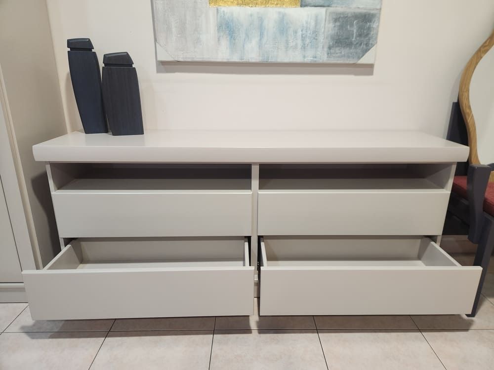 Una cómoda gris claro con dos columnas de cajones, las dos inferiores abiertas, colocada contra una pared blanca decorada con obras de arte.