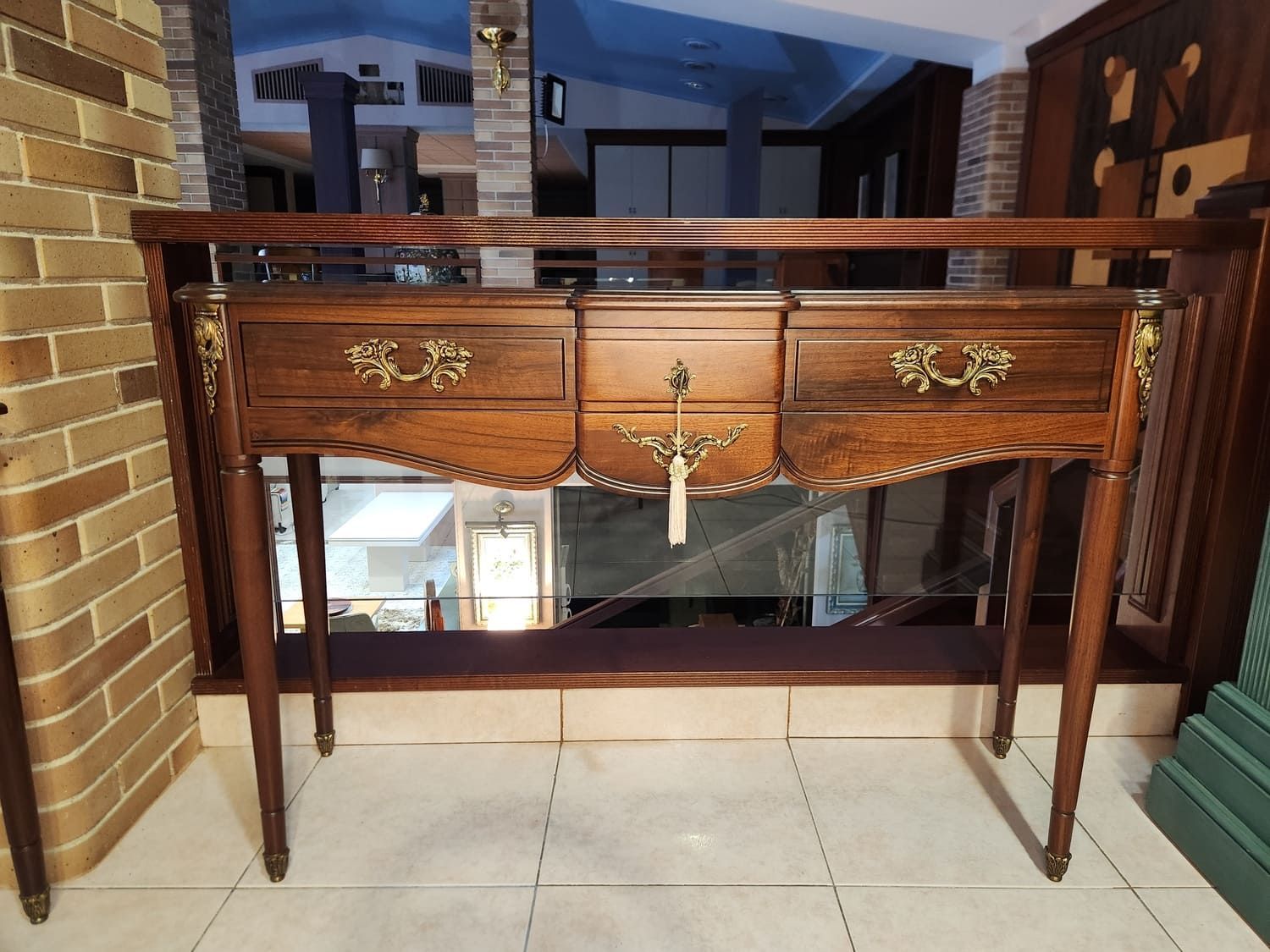 Consola de madera con cajones, herrajes dorados y patas delgadas contra una pared de ladrillos.