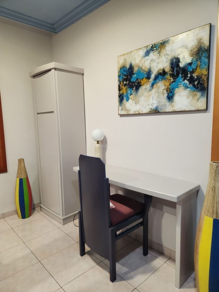 Un rincón de una habitación con un escritorio con una silla oscura, un armario alto, arte abstracto en la pared y jarrones de colores en el suelo.