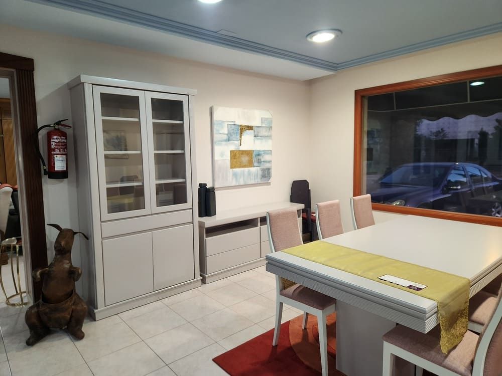 Un comedor con una vitrina gris, una mesa con un camino de mesa amarillo, sillas a juego y una ventana con vistas a un coche.