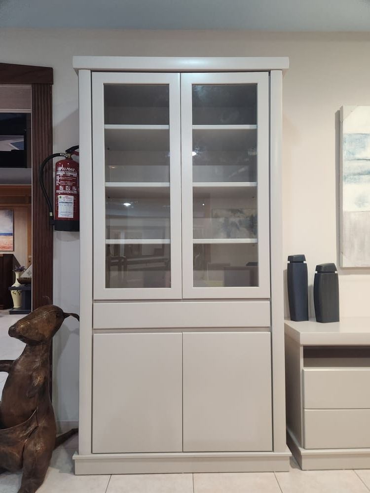 Un armario alto de madera gris claro con dos puertas superiores con paneles de vidrio y dos puertas inferiores macizas, situado en una habitación.