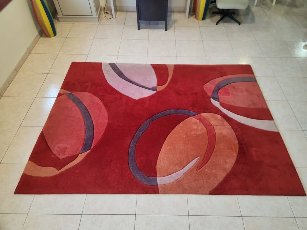 Una alfombra rectangular roja con formas circulares abstractas superpuestas en tonos rosas, naranjas y morados sobre un suelo de baldosas.