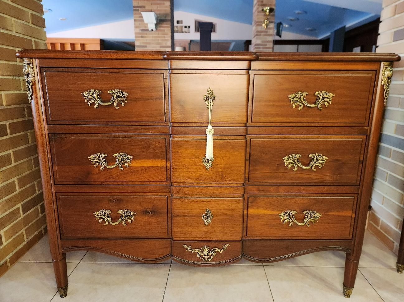 Cómoda de madera con nueve cajones, tiradores de latón ornamentados y base curva, colocada contra una pared de ladrillos.